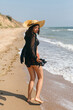 © sonyachny - Beautiful carefree woman with hat walking on sandy beach at sea waves and relaxing. Summer vacation. Fit stylish young female in light black shirt and straw hat enjoying vacation on tropical island