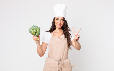 Wall Mural - pretty woman smiling and looking friendly, showing number two wearing an apron and holding a broccoli