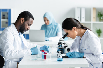 Wall Mural - Multiethnic scientists writing on clipboard and working with microscope in lab