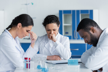 Wall Mural - Exhausted interracial scientist sitting near test tubes and clipboard