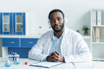 Wall Mural - African american scientist in headphones looking at camera near flasks and clipboard