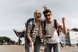 © Look! - Blonde woman in plaid shirt shoes v-sign. Travelers with backpacks rejoice. Brunette guy in white tee-shirt holds retro camera.