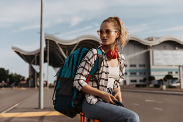 Tanned blonde woman in denim pants, white top and plaid shirt sits near airport. Tourist in sunglasses holds backpack.