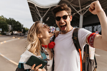 Brunette man happily screams and takes selfie with girlfriend near airport. Cool blonde girl holds passport and laughs.