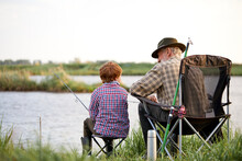 Rear View On Senior Man With Grandson Sitting Near River With Fishing Rods In Hands, Enjoying Beautiful Nature, Little Boy Learn To Fish With Grandfather. At Weekends In Evening, At Sunset
