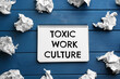 © New Africa - Notebook with text Toxic Work Culture and crumpled paper on blue wooden table, flat lay