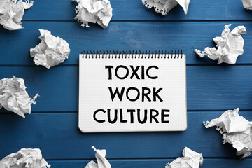 Notebook with text Toxic Work Culture and crumpled paper on blue wooden table, flat lay