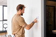 © Roman - Handsome man standing near wall-mounted device adjusting degrees in living room set comfortable temperature using thermostat home heating system. Owner of modern smart house, energy saving concept