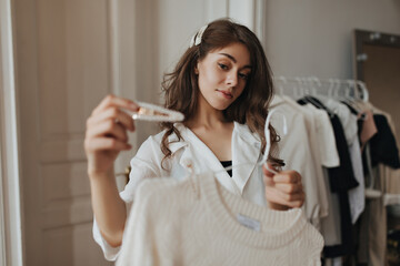 Pretty lady holding white hair pin and beige sweater. Brunette young woman in stylish blouse and black tee smiling and posing in cozy room