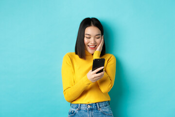 Wall Mural - Image of happy asian woman reading message on mobile phone screen and smiling, chat in smartphone app, standing over blue background