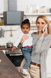 happy mother talking on smartphone and holding adopted african american kid in kitchen