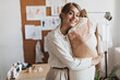 © Look! - Happy woman smiling and hugging mannequin. Joyful brunette girl in beige blouse and pants loves her job as fashion designer and poses in office