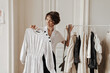 Portrait of curly short-haired woman in light blouse and black skirt poses in dressing room and holding hanger with white dress.