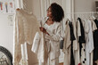 Cute curly dark-skinned woman in stylish pants and jacket poses in dressing room and looks at trendy knitted sweater.