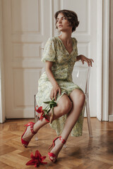 Wall Mural - Beautiful young brunette woman in stylish floral dress and red high heels sits on transparent chair and holds flowers.