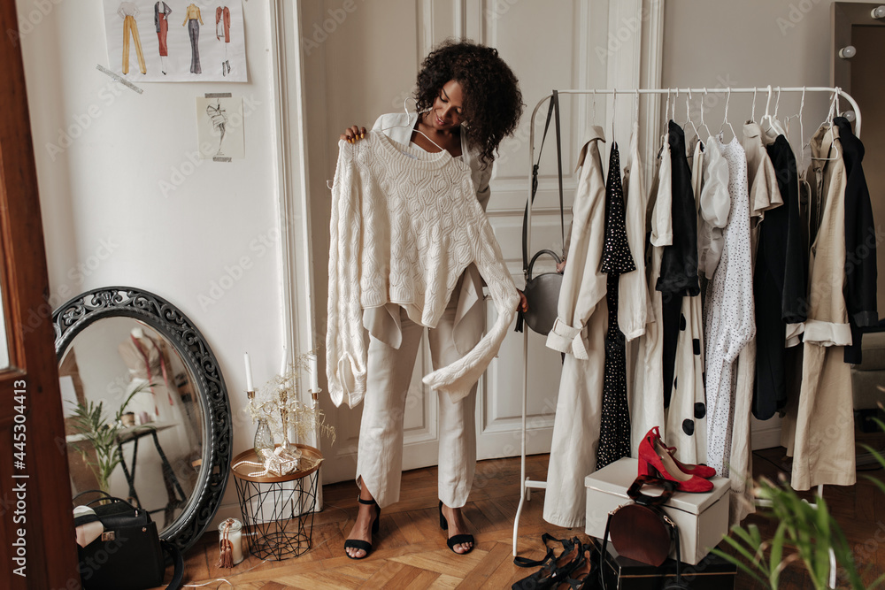 Stylish young dark-skinned curly lady in beige pants and jacket smiles, poses in dressing room an holds hanger with trendy knitted sweater. - obrazy, fototapety, plakaty 
