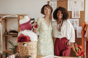 Wall Mural - Cheerful attractive ladies in stylish outfits smile and work as fashion designers. Woman in floral dress holds textile samples.