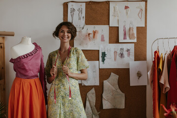 Wall Mural - Attractive brunette short-haired woman in floral yellow dress smiling sincerely, holding measuring tape and posing near mannequin.