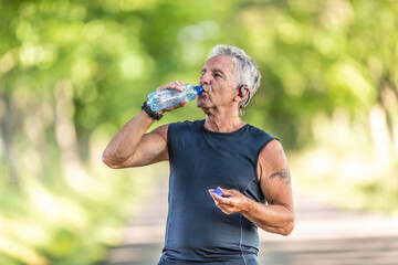 Wall Mural - Handsome elderly man rehydrates after a run outdoors in the nature by drinking water from a bottle