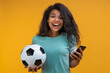 © wpadington - Studio portrait of  smiling lucky football fan girl holding ball in her hand and making bets on favourite team at bookmaker's website using smartphone