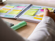 © OlgaPS - Close-up of agenda organize with color-coding sticky notes for time management. Productive schedule for appointments and reminders. Hand holding a yellow highlighter marker. Organization and planning
