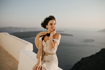 Wall Mural - Brunette short-haired woman in elegant beige dress and boater enjoys beautiful sea view. Curly lady in good mood sits on white fence.