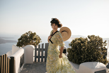 Sticker - Charming woman in floral dress looks into camera outside. Brunette lady in summer outfit holds boater and moves on sea background.