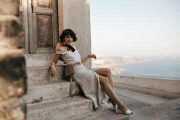 Wall Mural - Short-haired brunette woman in midi dress looks into camera and sits on stairs near old house with sea view. Charming lady in summer outfit poses near wooden door.