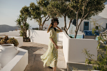 Wall Mural - Pretty brunette woman in midi dress and hat looks into camera and smiles. Lady in floral outfit walks in old Greek city.