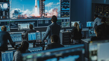 Group Of People In Mission Control Center Witness Successful Space Rocket Launch. Flight Control Employees Sit In Front Computer Displays And Monitor The Crewed Mission. Team Stand Up And Watch.