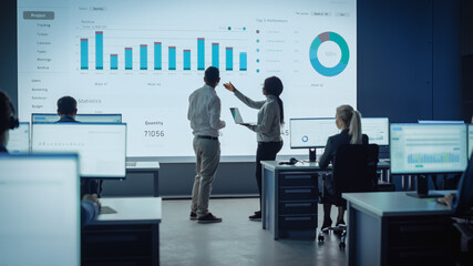 Two Traders Having a Meeting in a Modern Monitoring Office with Analytics Feed on a Big Digital Screen. Monitoring Room with Brokers and Finance Specialists Sit in Front of Computers. Colleagues Talk.