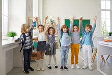 Group Portrait Of Happy Elementary School Students. Bunch Of Cute Primary School Children In Casual Wear With School Bags Standing In A Modern Classroom And Raising Their Hands Up