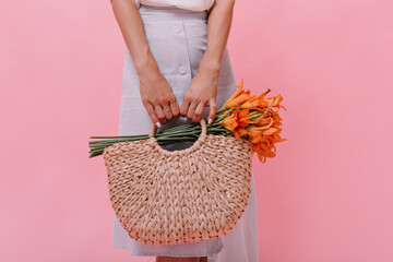 Lady poses with knitted bag and flowers on pink background. Woman in blue light skirt holds straw handbag with orange beautiful bouquet