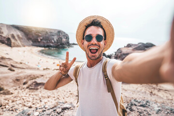 Wall Mural - Young man with backpack taking selfie portrait on a mountain - Smiling happy guy enjoying summer holidays at the beach - Millennial showing victory hands symbol to the camera - Youth and journey