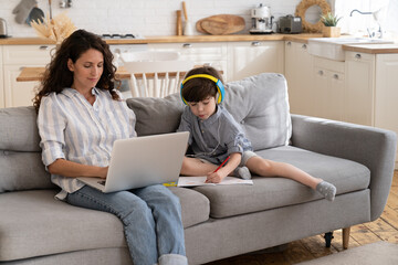 Wall Mural - Casual businesswoman work online on laptop computer as little son write in textbook listen to music or audio lesson from preschool education courses. Home school and remote office during coronavirus