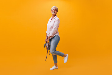 Wall Mural - Charming woman in jeans, shirt and sunglasses poses on orange background