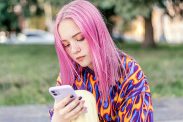 Pink-haired teenage hipster girl in a colorful bright T-shirt is using a smartphone on a summer day.Summer concept.Generation Z style.Social media concept.