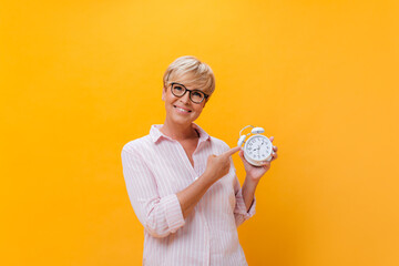 Wall Mural - Beautiful lady in eyeglasses looks into camera and points to alarm clock on orange background