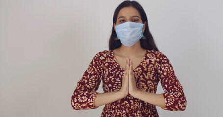 Poster - Young Indian woman with a mask gesturing Namaste or Vanakkam on a white wall background