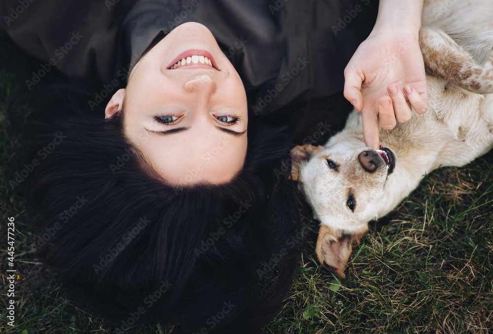 A young cheerful girl looks at the viewer and lies on the green grass near a mongrel dog, which playfully bites her finger. The concept of friendship between man and animal. - obrazy, fototapety, plakaty 