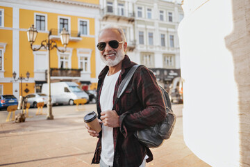 Charming man in sunglasses holds coffee cup. Attractive adult guy in light T-shirt and burgundy shirt posing with backpack..