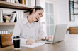 © topkritsada - Pretty businesswoman sitting on the phone at her desk with laptop computer and notepad in hustle and bustle, rush hour work concept, working from the smart office.