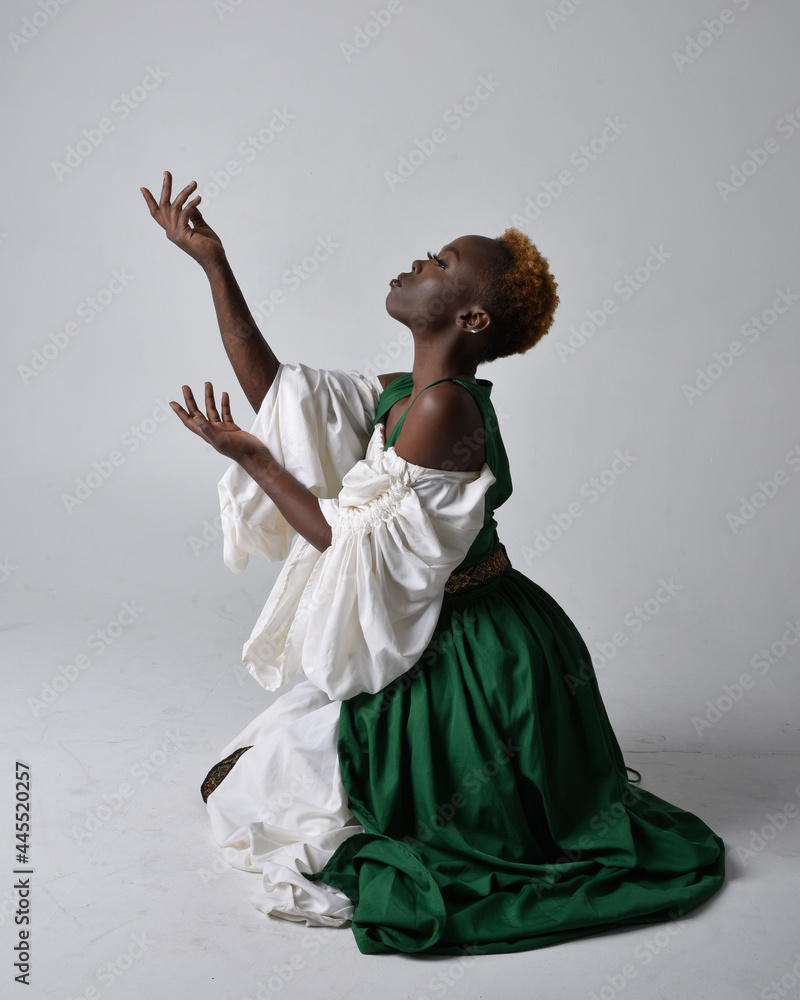 Full length portrait of pretty African woman wearing long green medieval  fantasy gown, kneeling pose on a light grey studio background. Stock Photo  | Adobe Stock