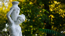 Sculpture Of A Girl In The Spring Park. An Old Statue In A Park Of A Sensual Semi-nude Greek Or Italian Renaissance Woman With A Vase In A City Park. Sunny Day In The Summer Garden. Close-up