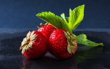Fresh Ripe Strawberries Or Fragaria Ananassa On A Dark Background. Three Red Berries Decorated With Fresh Semi-transparent Mint Leaves. Diet, Natural Products, Harvest, Nutrition, Food Decorations.