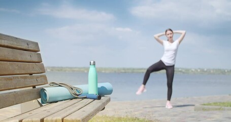 Poster - Sporty young woman training near river, focus on jumping rope, yoga mat and bottle of water on bench