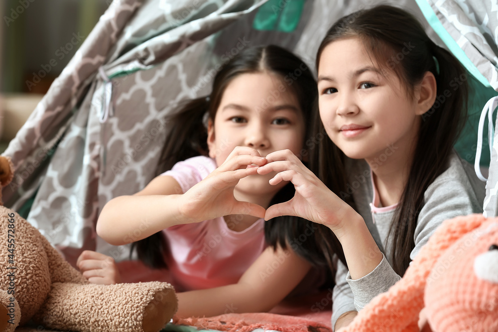 Cute Asian sisters making heart shape with their hands at home