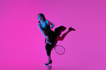 Wall Mural - High-fashion styled young woman in black evening gown, dress playing tennis isolated over pink neon background. Concept of fashion and sport