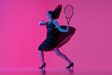 Wall Mural - High-fashion styled young woman in black evening gown, dress playing tennis isolated over pink neon background. Concept of fashion and sport