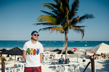 Young tourist man with sunglasses smiling and having good time at the beach, posing cheerful against blue sky and palm tree